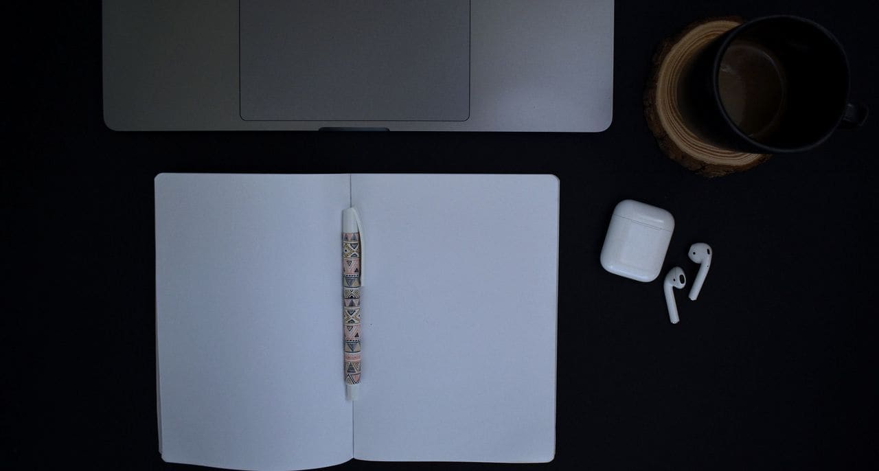A laptop, open notebook and coffee on a black desk, seen from above